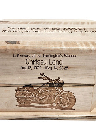 A wooden urn or casket with a carved design of a motorcycle on it. The urn has a message engraved on it, which reads "The best part of any journey is the people we meet along the way" and "In Memory of our Huntington's Warrior Chrissu Land July 12, 1972 - May 14, 2035".