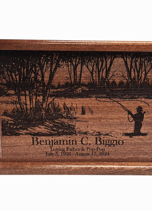 a wooden urn or urn-like container with a carved design depicting a person fishing in a wooded area. The text on the container reads "Benjamin C. Biggio Living Father & Son July 5, 1956 - August 17, 1964".